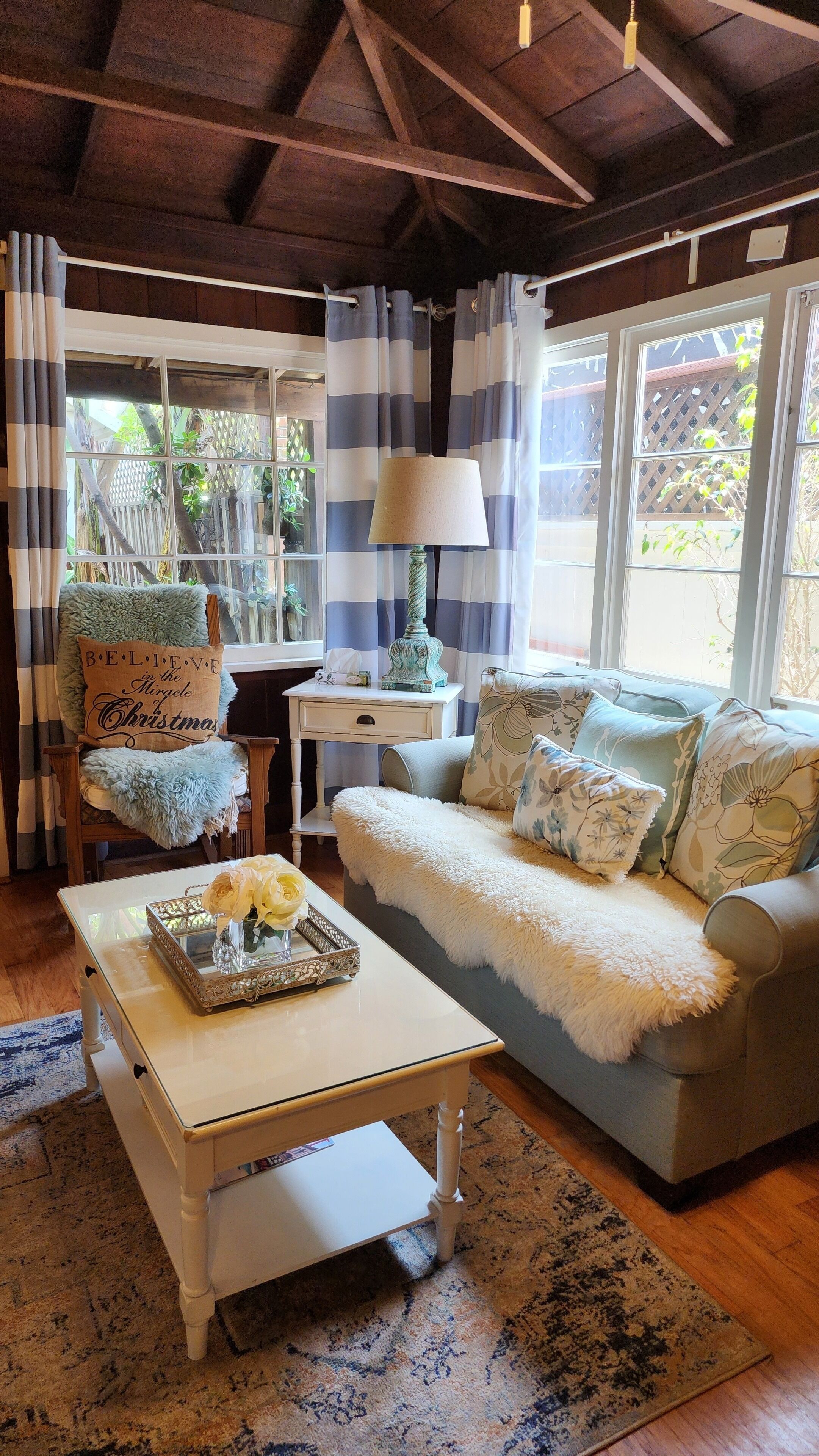 A cozy living room with a wooden ceiling, a sofa with floral cushions, a white coffee table with a vase of flowers, and a striped curtain.