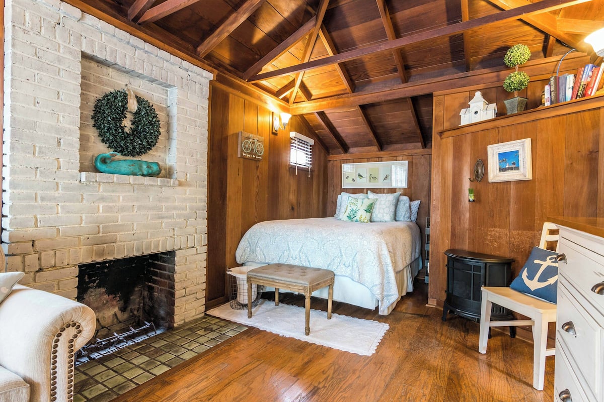 Una habitación acogedora con chimenea de ladrillo, una cama con edredón azul y blanco, vigas de madera en el techo y una pequeña mesa con una silla.