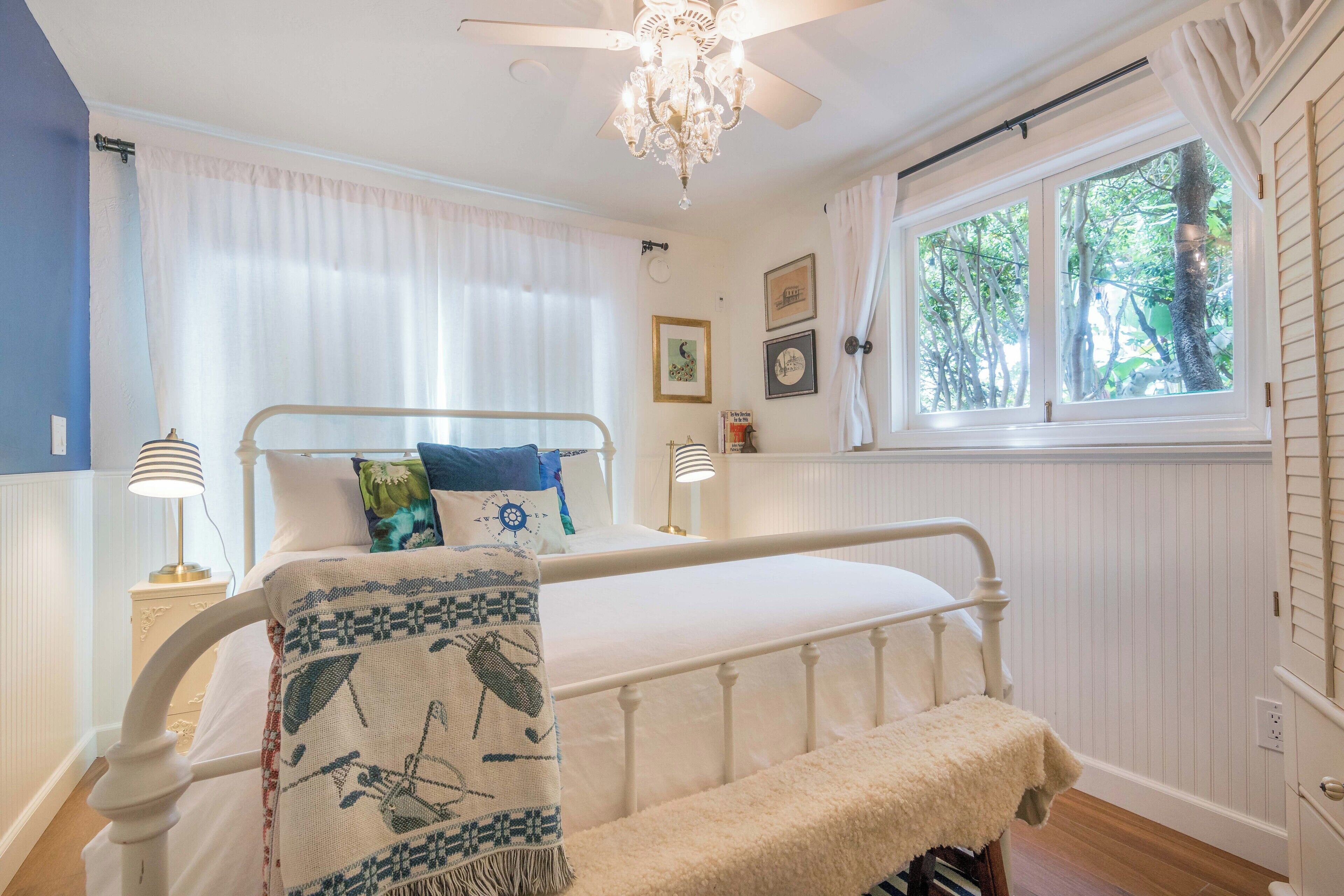 Une chambre à coucher avec un lit blanc, un banc, un lustre, une fenêtre donnant sur des arbres et des lampes fixées au mur.