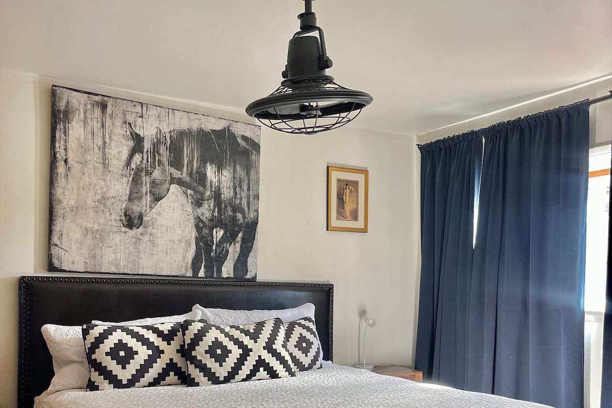 Un dormitorio con una cama grande, un ventilador de techo, un cuadro de un caballo en la pared, una fotografía enmarco y una ventana con cortinas.