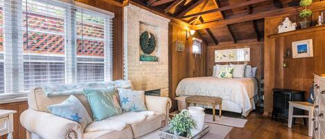 Una habitación acogedora con techo de madera, una cama, un sofá blanco con almohadas de estrellas, una mesita de centro blanca con una planta y una chimenea.