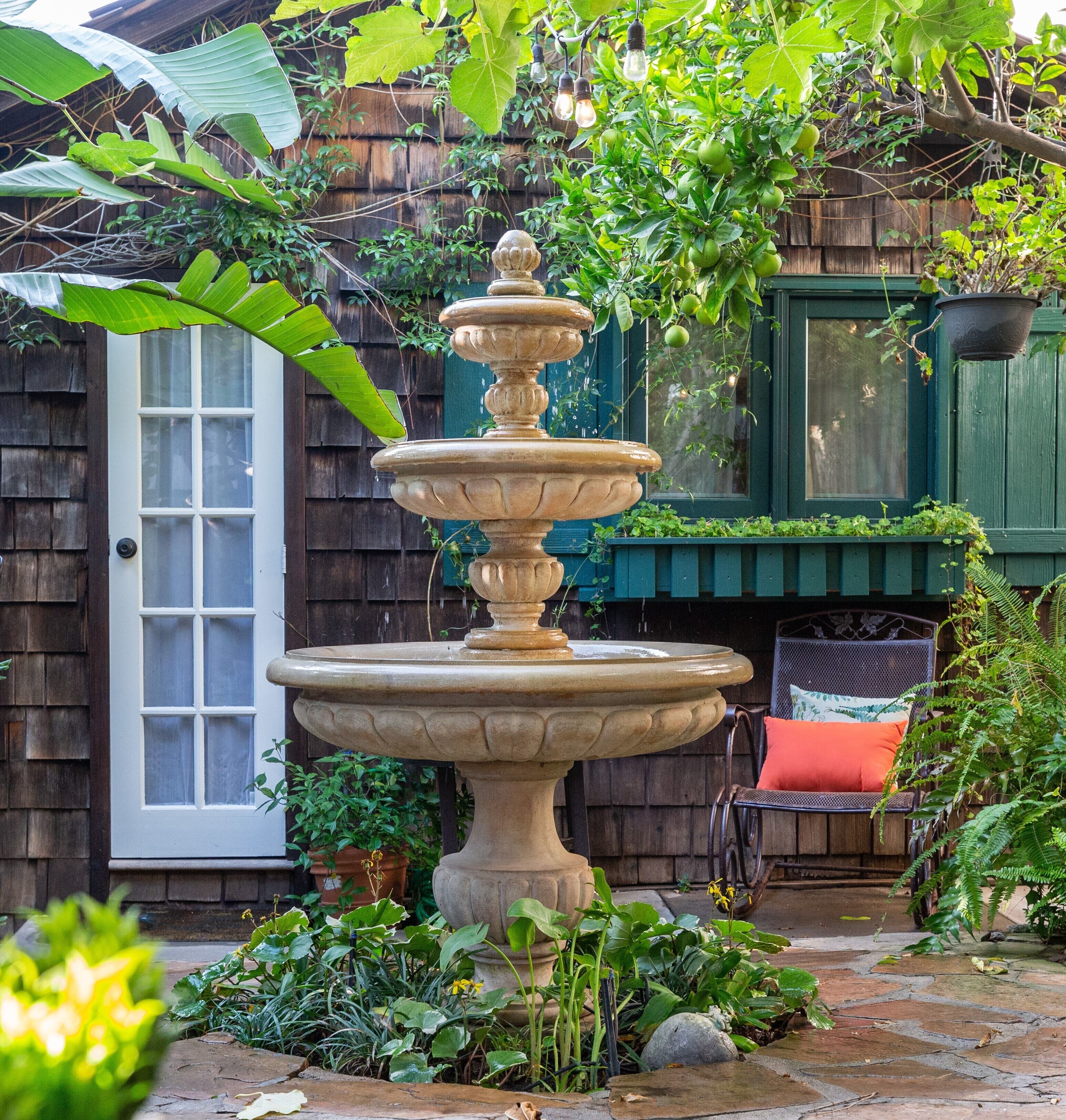 A tiered stone fountain in a garden with a wooden door and a chair with a cushion.