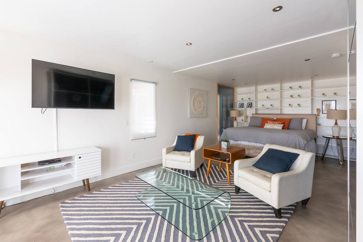 Un dormitorio moderno con una cama, dos sillones, una mesa de centro de vidrio, un televisor y estanterías empotradas.
