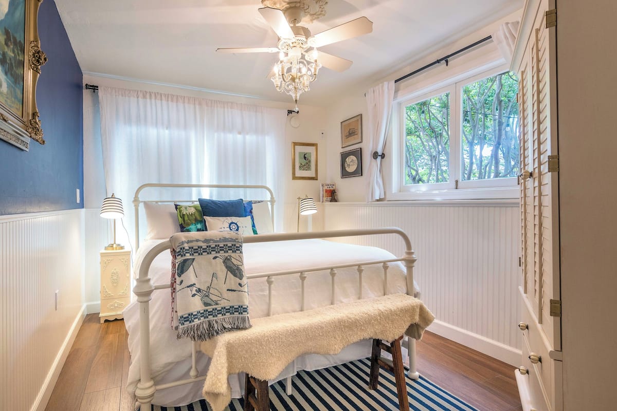 Un dormitorio con una cama de metal blanco, un ventilador de techo, una araña y una ventana con cortinas blancas, con vistas a árboles.