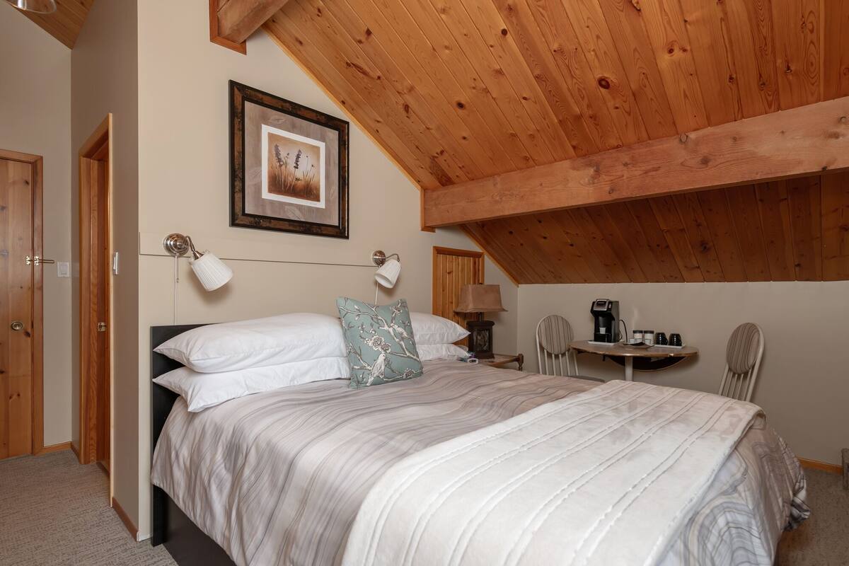 A bedroom with a wooden ceiling, a bed with white bedding, a framed picture on the wall, and a small table with a coffee maker.
