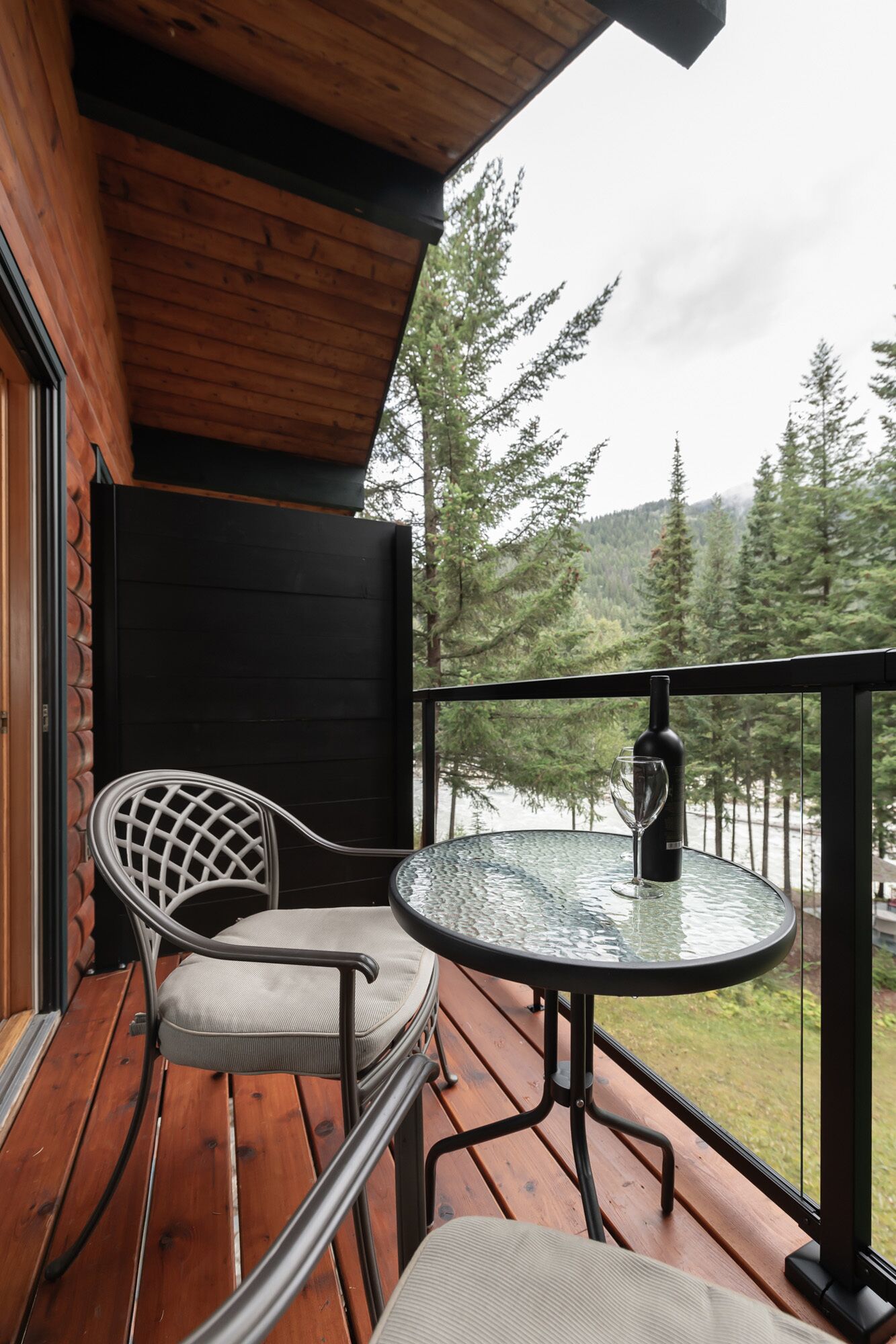 A wooden deck with a glass table and metal chair, overlooking a forested area.