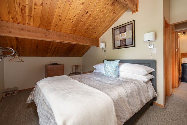 Un dormitorio con techo de madera, una cama con ropa blanca, un aparador, un espejo y un cuadro en la pared.