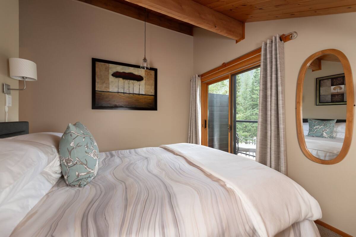 A bedroom with a bed, a framed painting, a window with curtains, and a mirror on the wall.