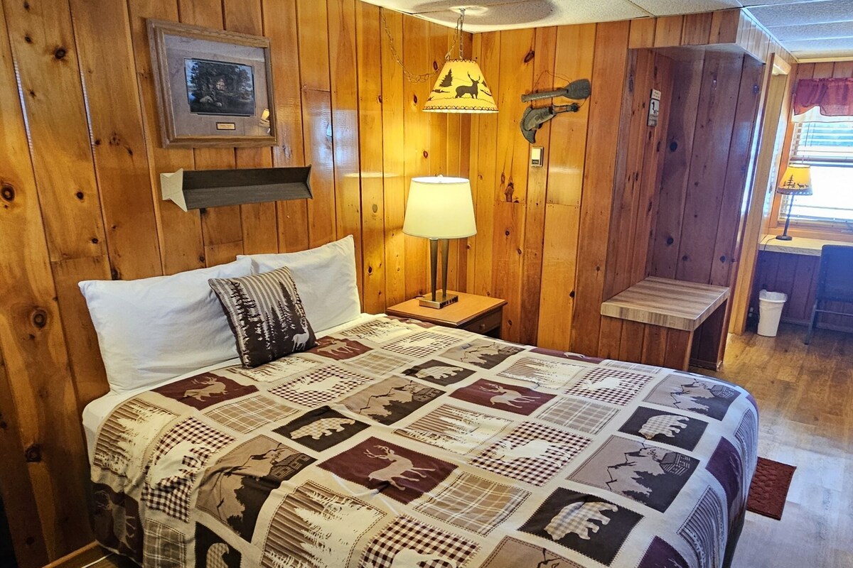 Een kamer met houten lambrisering, een bed, nachtkastje, lamp, een klein tafeltje en een raam met jaloezieën.