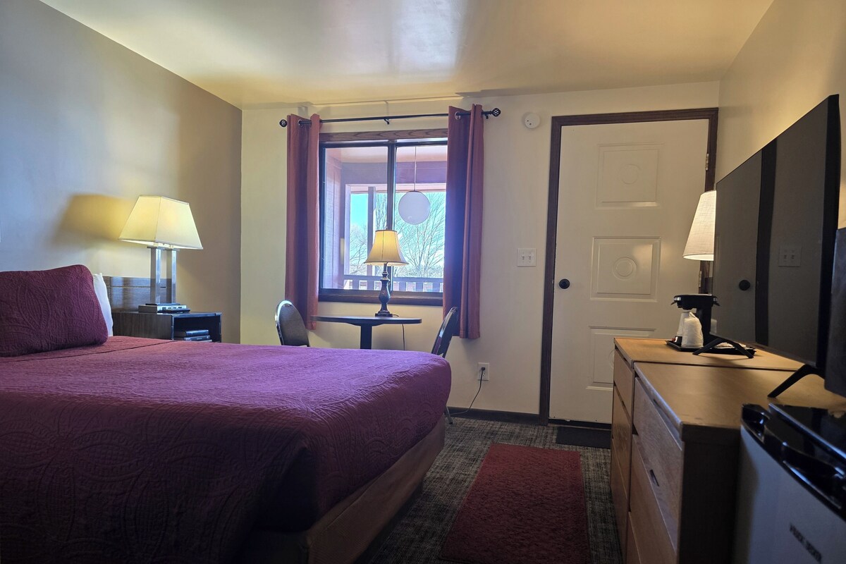 Hotelkamer met een groot bed, een bureau, een stoel, een klein tafeltje, een lamp, een koffiezetapparaat en een deur.