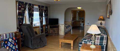 A living room with a sofa, armchair, wooden coffee table, and a television. There is a kitchen visible through an archway.
