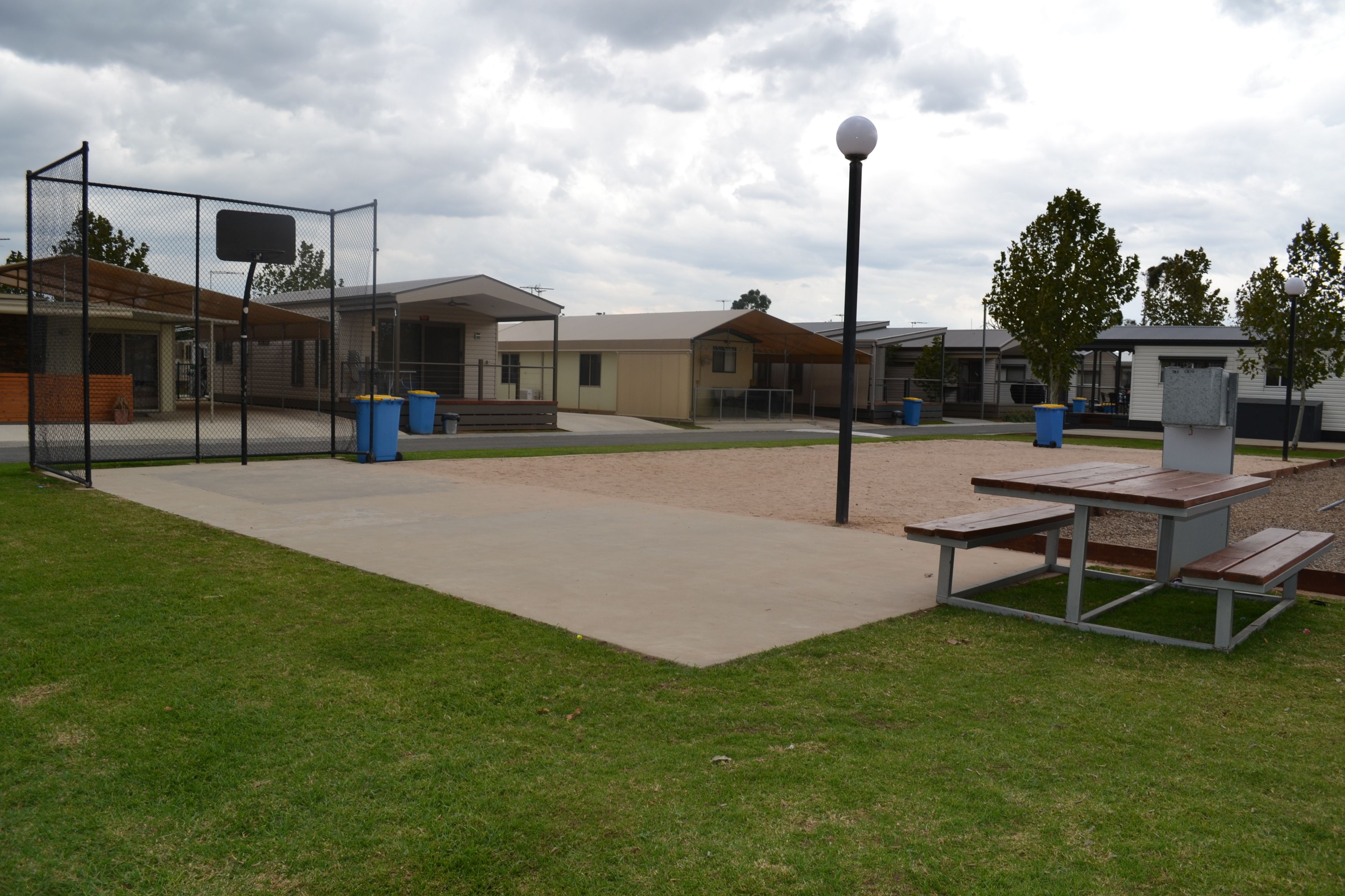 An outdoor recreational area with a basketball court, picnic tables, trash bins, and a covered seating area.