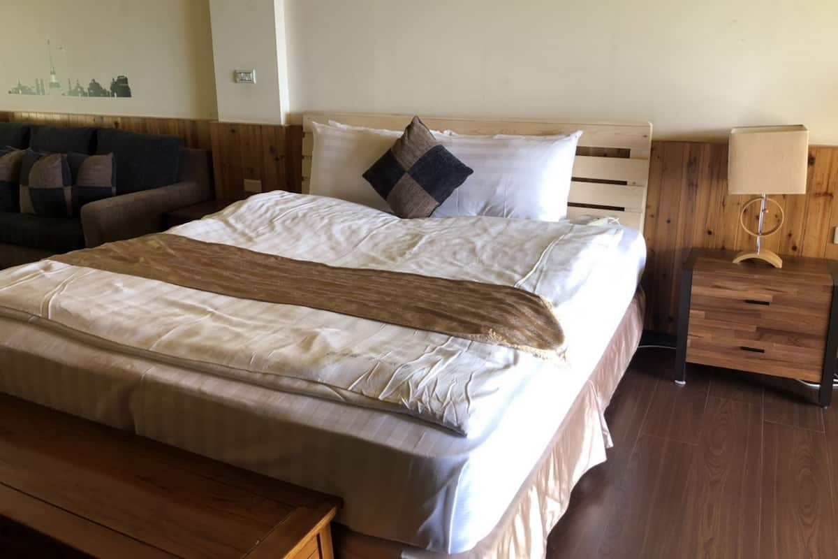 Ein ordentlich bezogenes Bett mit einem hölzernen Kopfteil, ein Nachttisch mit Lampe und eine Holzbank.