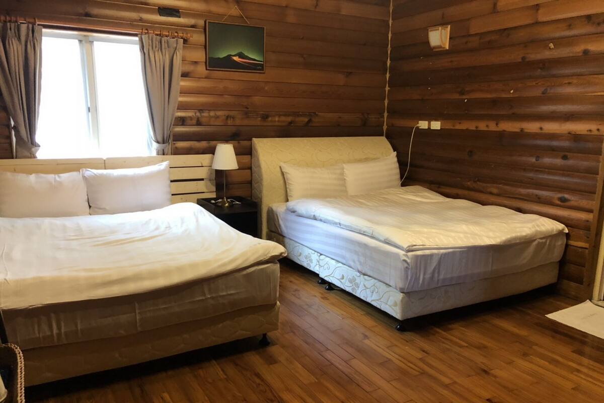 Ein Zimmer einer Holzhütte mit zwei Betten, einem Nachttisch, einer Lampe und einem Fenster mit Vorhängen.