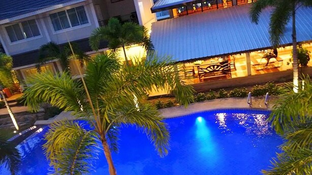 A swimming pool surrounded by palm trees and a building with a covered patio area. - Good Times Resort (Kanchanaburi)