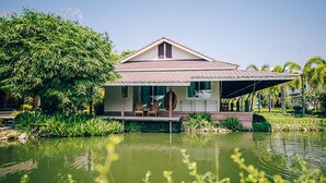 A house with a tiled roof, a covered porch, and a pond in the foreground. - Good Times Resort (Kanchanaburi)