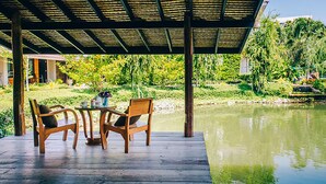 A wooden deck with two chairs and a table, overlooking a pond and lush greenery. - Good Times Resort (Kanchanaburi)