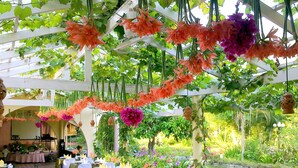 Un área de comedor al aire libre con mesas dispuestas para una comida, rodeada de abundante follaje y flores colgantes.