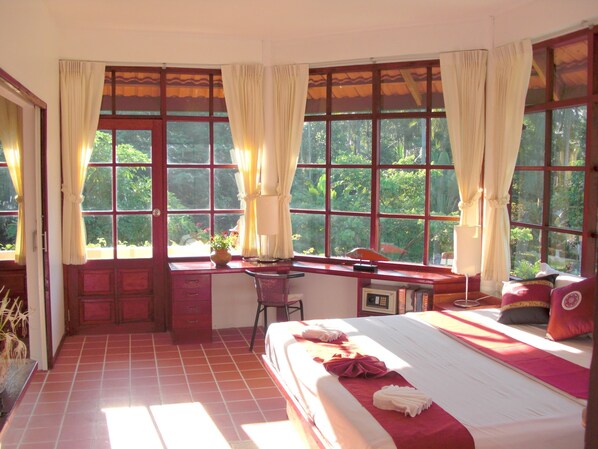 A room with a bed, a desk, and a view of greenery through large windows. - Ekman Garden Resort Nakhon Si Thammarat (Sichon)