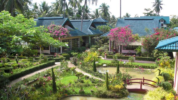 Un jardín exuberante con un estanque, palmeras y edificios de estilo tradicional.
