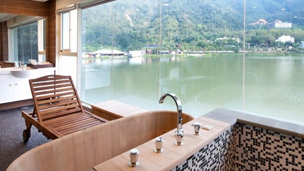 A modern bathroom with a wooden bathtub, a large window offering a view of a lake and mountains, and a wooden chair. - Lakeside Resort Hotel (Puli)