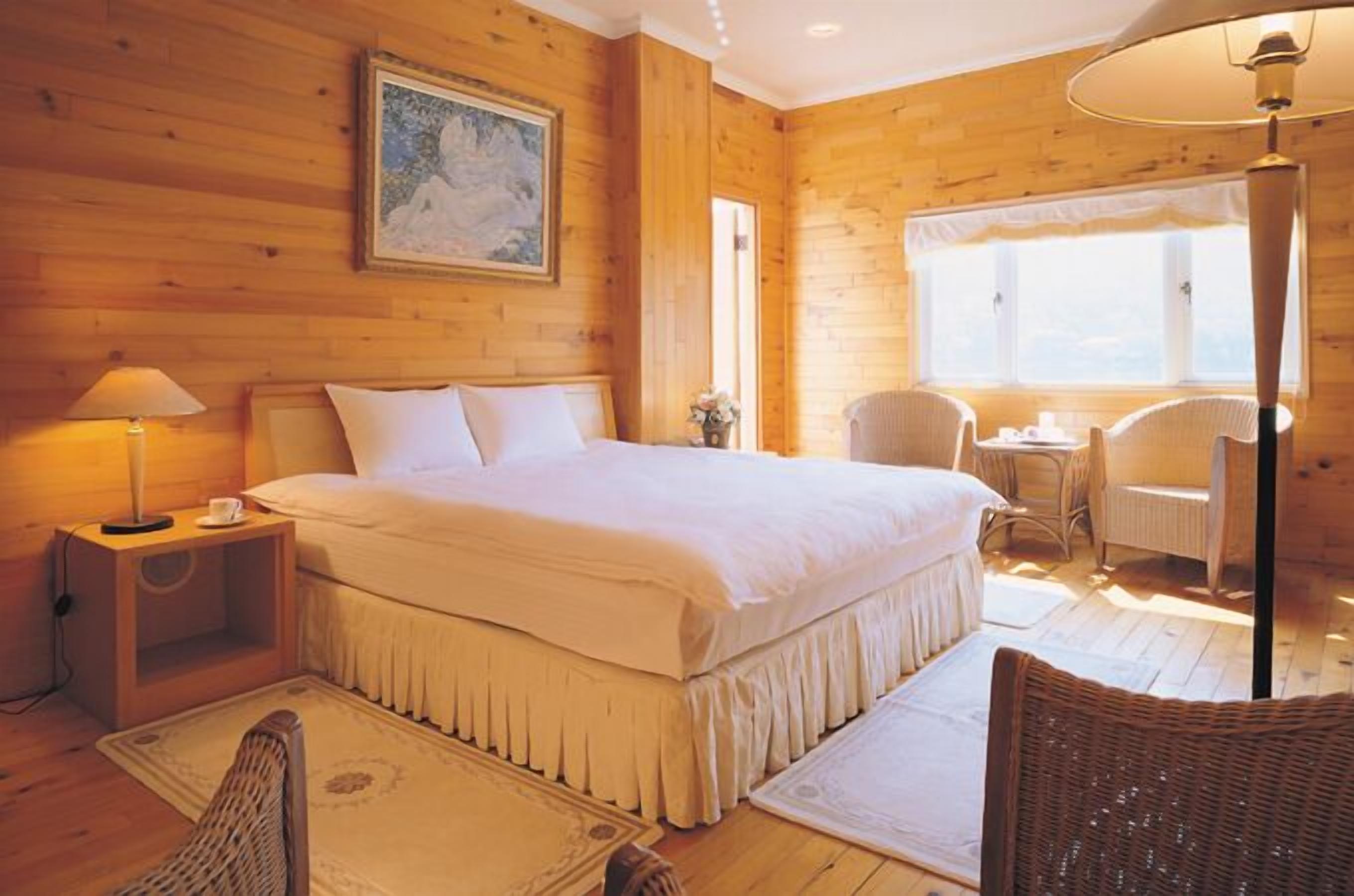 A wooden room with a bed, a nightstand, a lamp, a chair, and a window.