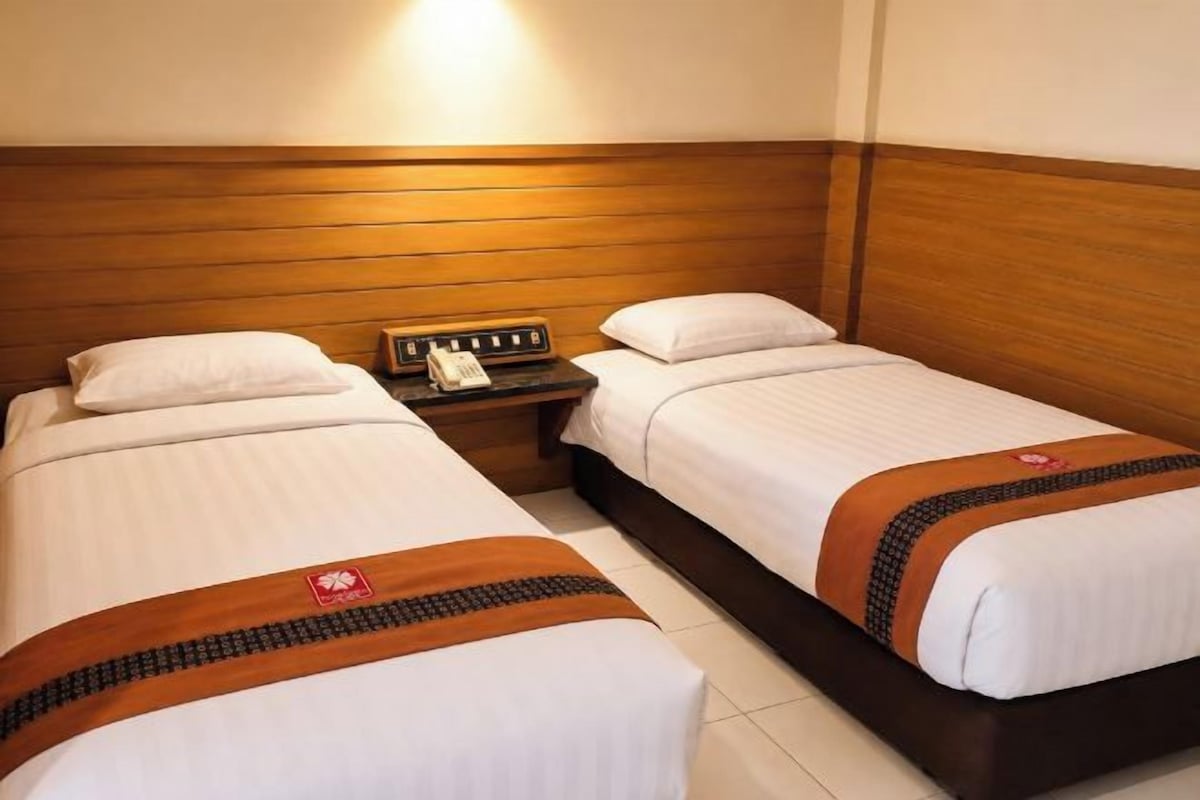 Habitación de hotel con dos camas individuales, un cabecero de madera, una mesita de noche con teléfono e iluminación empotrada en la pared.