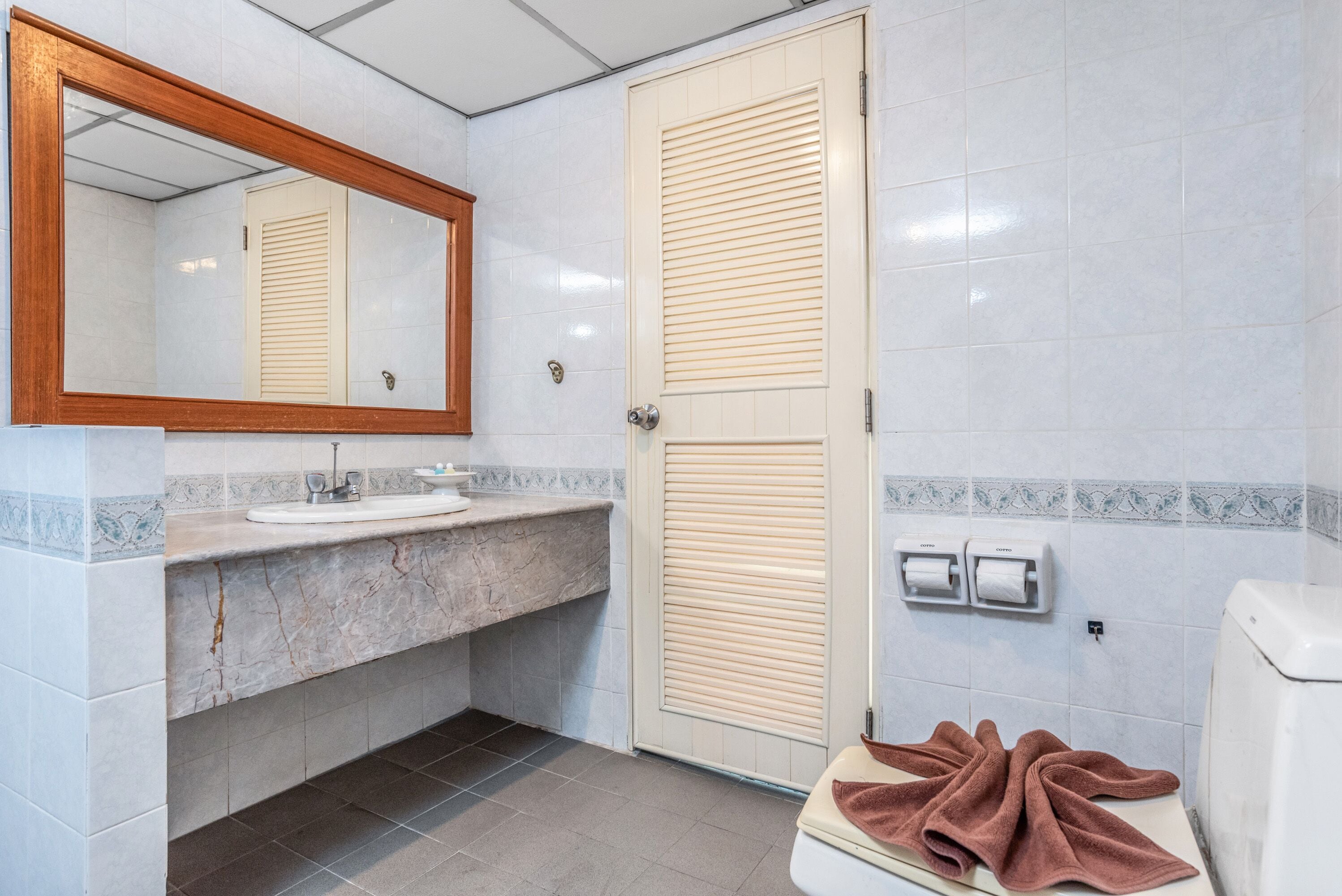 Un baño con lavamanos de mármol, espejo con marco de madera, inodoro con toalla y puerta con panel persiana.