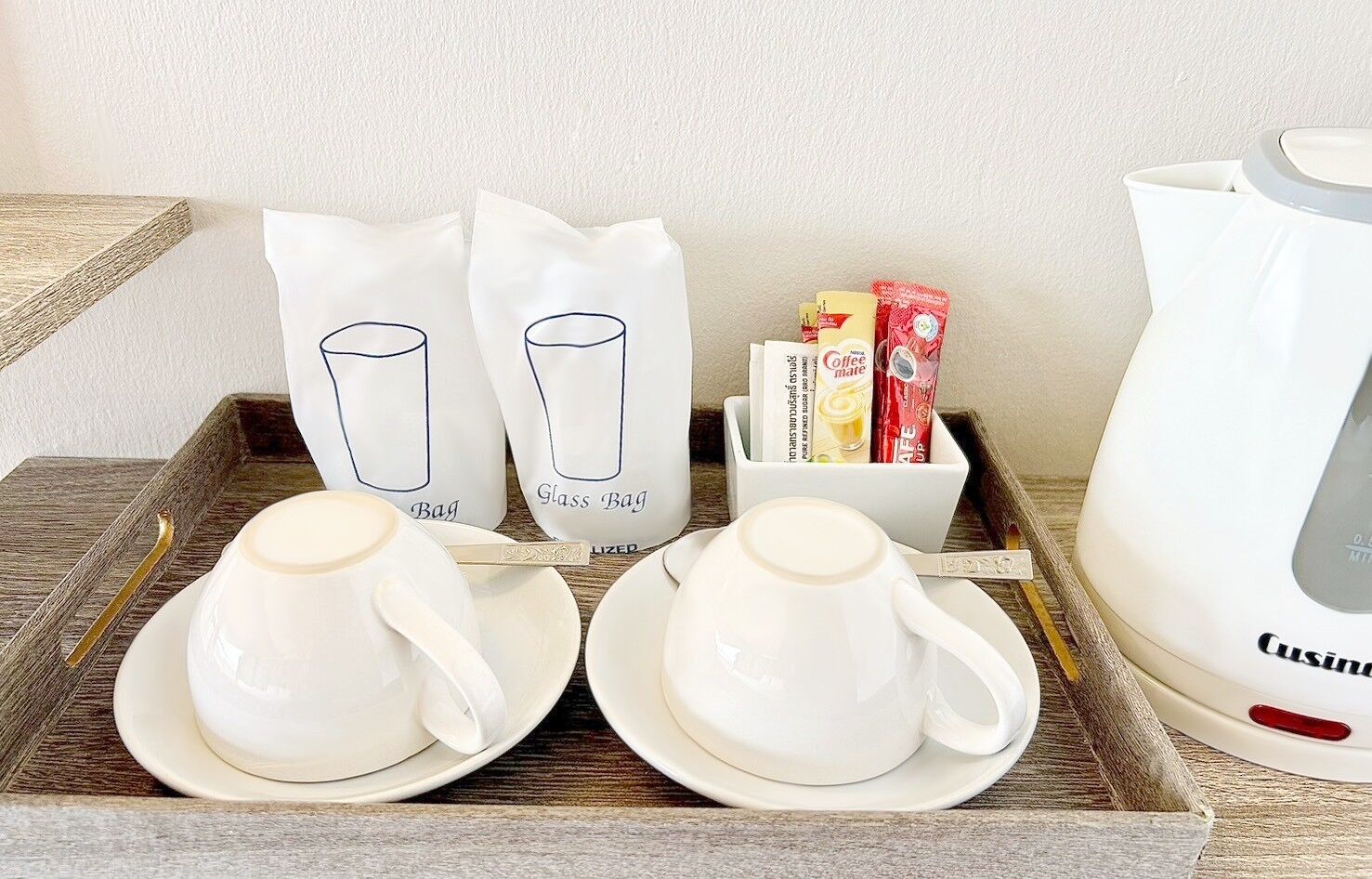 Een dienblad met twee witte glazen kopjes en schotels, twee witte zakken met de tekst 'Glass Bag', een witte koffiezetapparaat en een witte verpakking met koffiehoeveelheden.