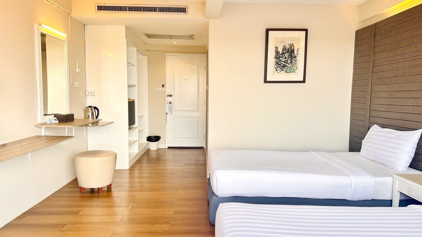 Una camera d'albergo moderna con pavimento in legno, un letto bianco, un poggiapiedi rotondo e un comodino bianco, e un quadro appeso al muro.