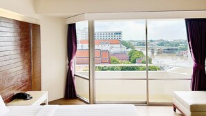 A hotel room with a large window, a bed, a bedside table with a phone, and a view of buildings and a bridge. - Ayothaya Riverside Hotel Ayutthaya (Ayutthaya)