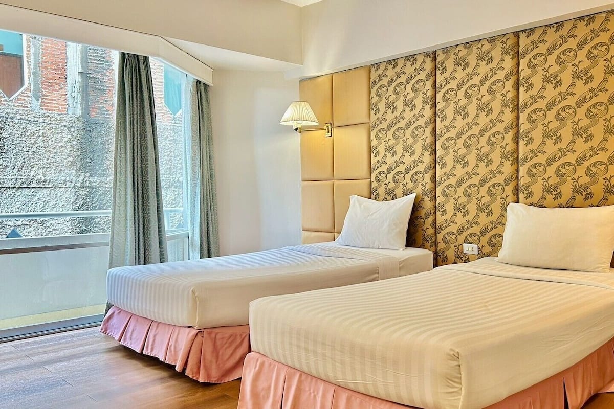 A hotel room with two beds, a large window with curtains, a patterned headboard, and a bedside table with a lamp.