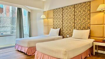 A hotel room with two beds, a large window with curtains, a patterned headboard, and a bedside table with a lamp.