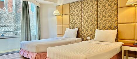 A hotel room with two beds, a large window with curtains, a patterned headboard, and a bedside table with a lamp.