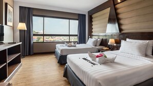 Ein Hotelzimmer mit zwei Betten, einem Schreibtisch, einer Lampe und einem großen Fenster mit Blick auf die Stadt.