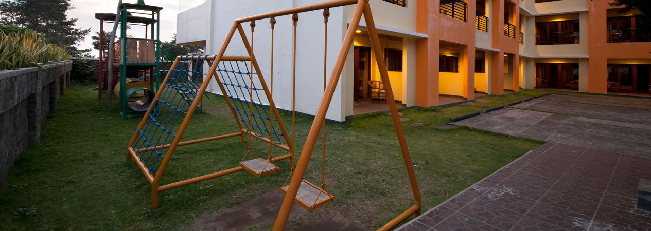 A playground with swings and a climbing structure, adjacent to a building with multiple balconies and a paved courtyard.