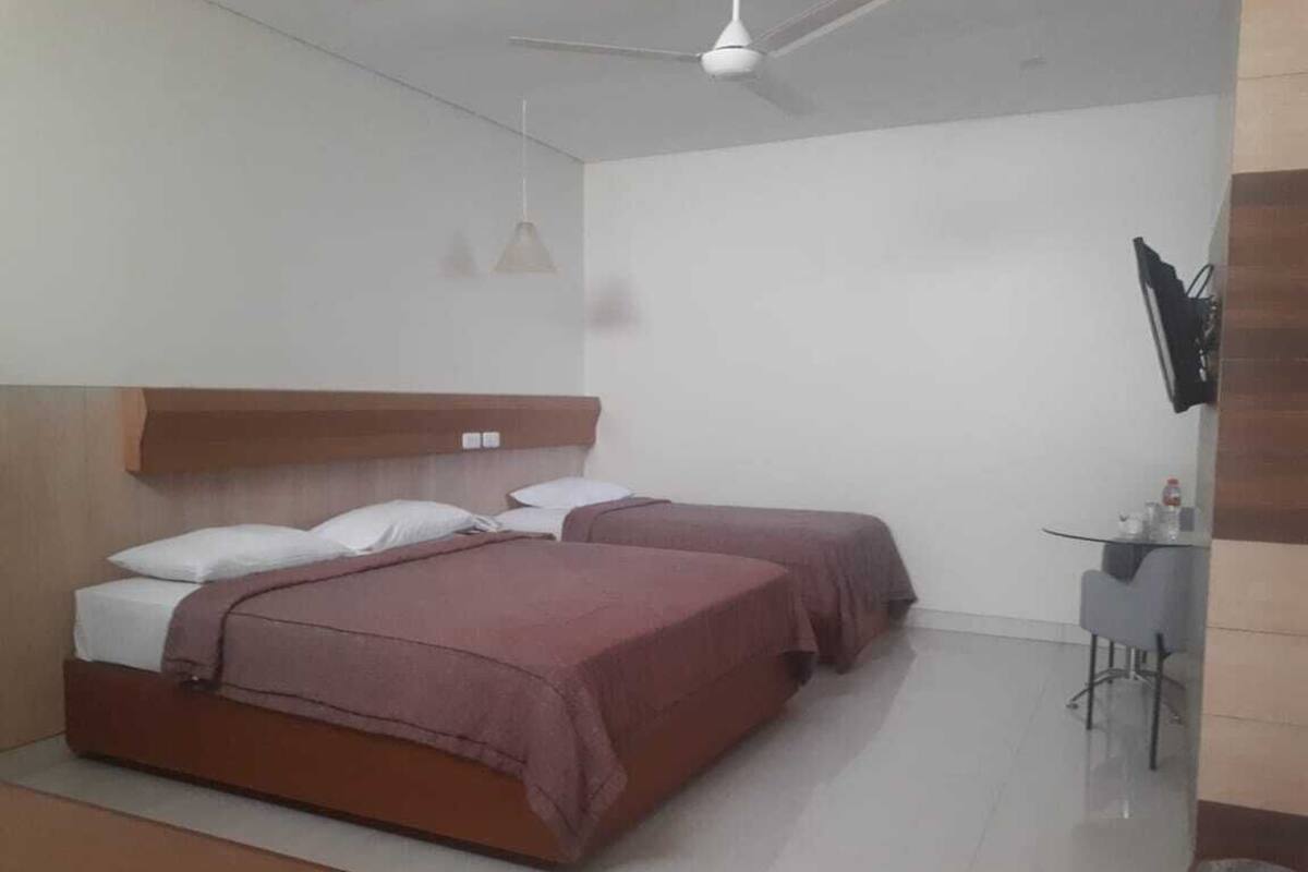 A hotel room with two beds, a small table, a TV mounted on the wall, and a ceiling fan.