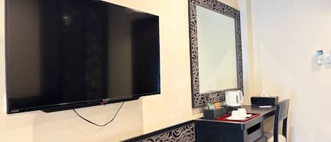 Habitación de hotel con televisor de pantalla plana montado en la pared, un espejo decorativo con marco ornamentado y una mesita con hervidor y pañuelos.