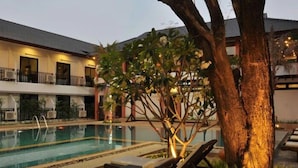 Piscina com espreguiçadeiras e uma árvore, cercada por um edifício com varandas.