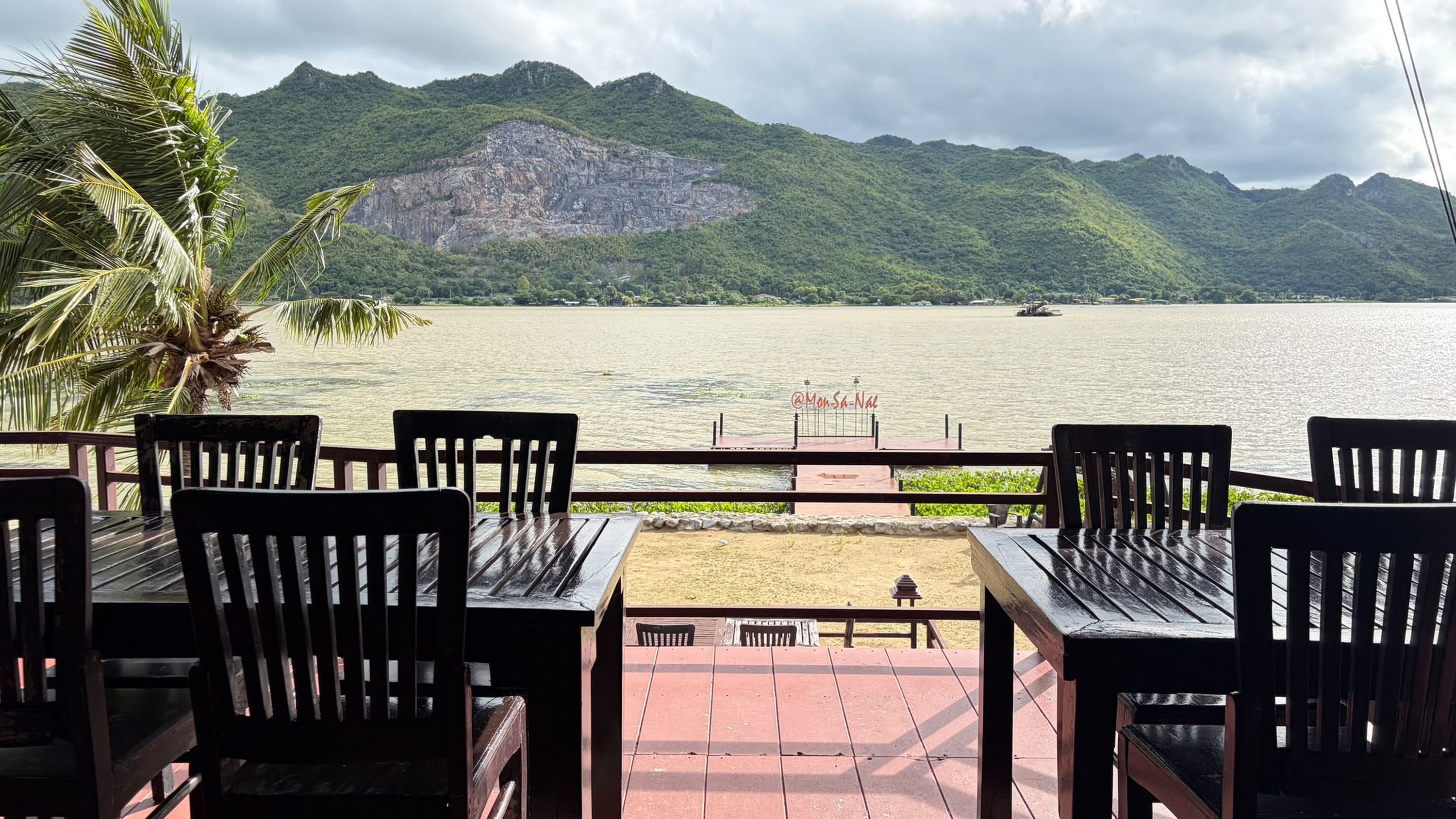 Una terraza con mesas y sillas de madera, con vistas a un cuerpo de agua y montañas al fondo.