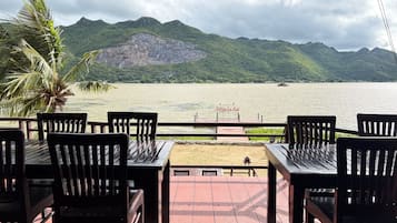 Una terraza con mesas y sillas de madera, con vistas a un cuerpo de agua y montañas al fondo.
