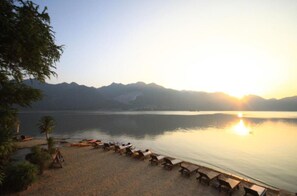 A beach with a row of sun loungers and a calm lake, surrounded by mountains. - Palette Monsane River Kwai Resort (Kanchanaburi)