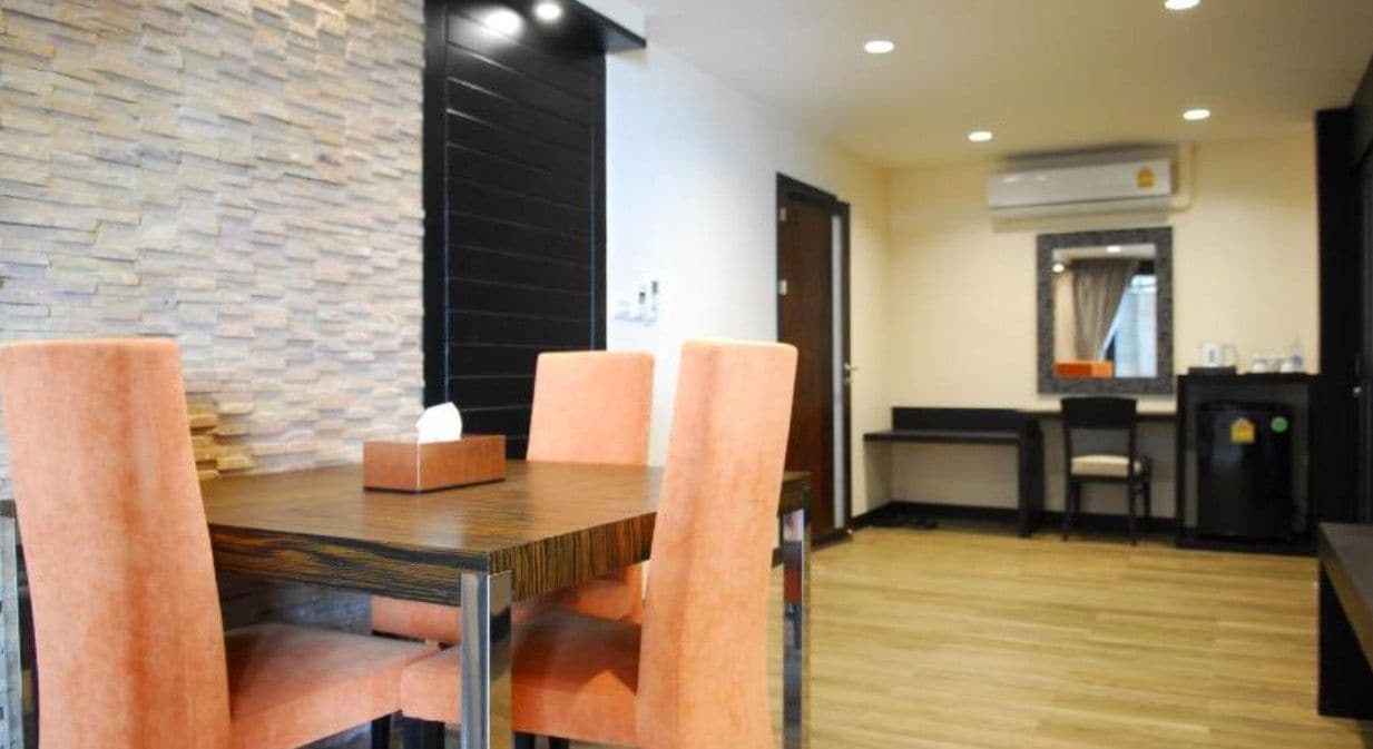 A dining area with a wooden table and four chairs, a stone wall, and a small kitchen area with a refrigerator and a sink.