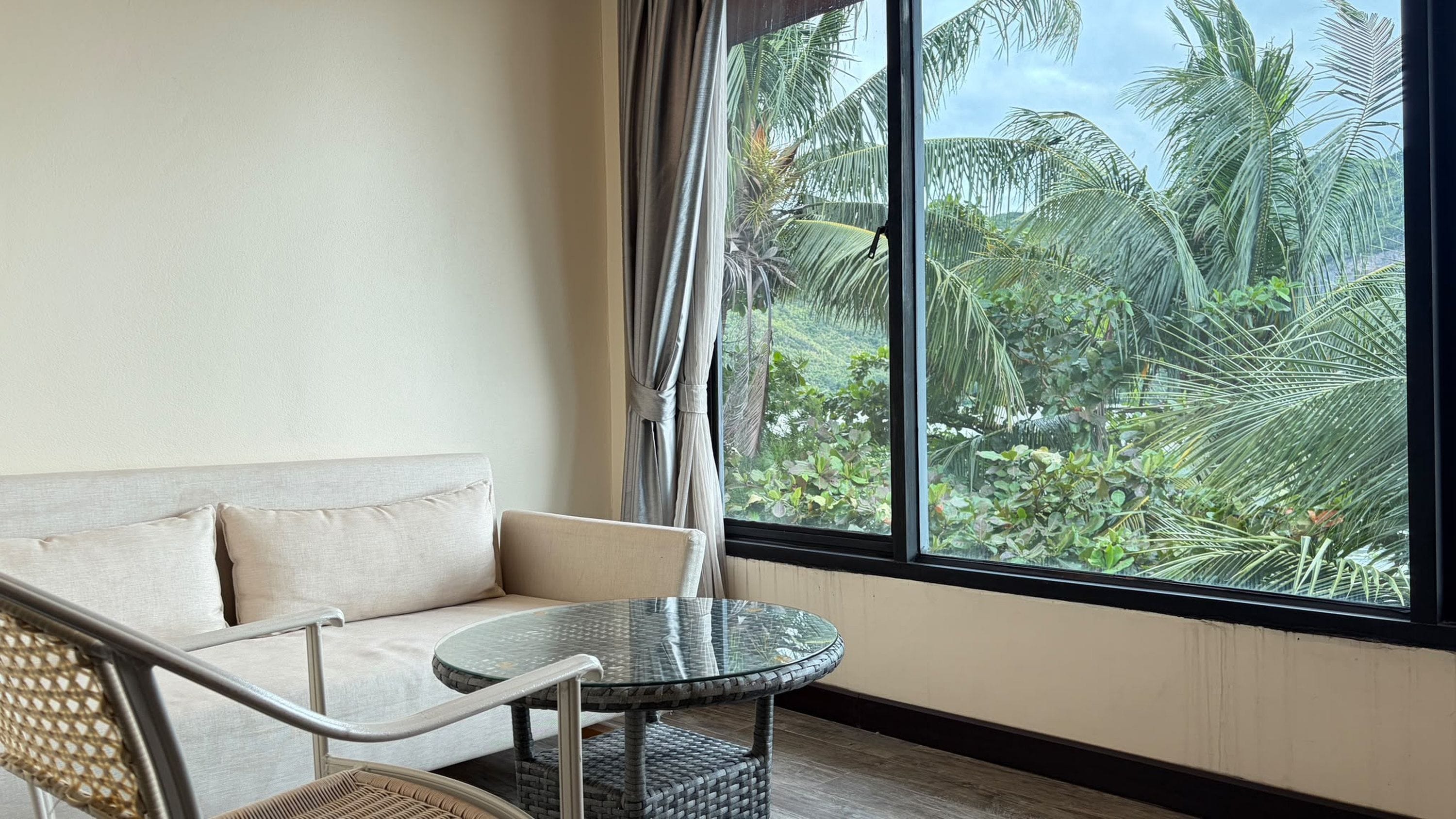 A room with a sofa, a round glass table, and a view of tropical foliage through large windows.