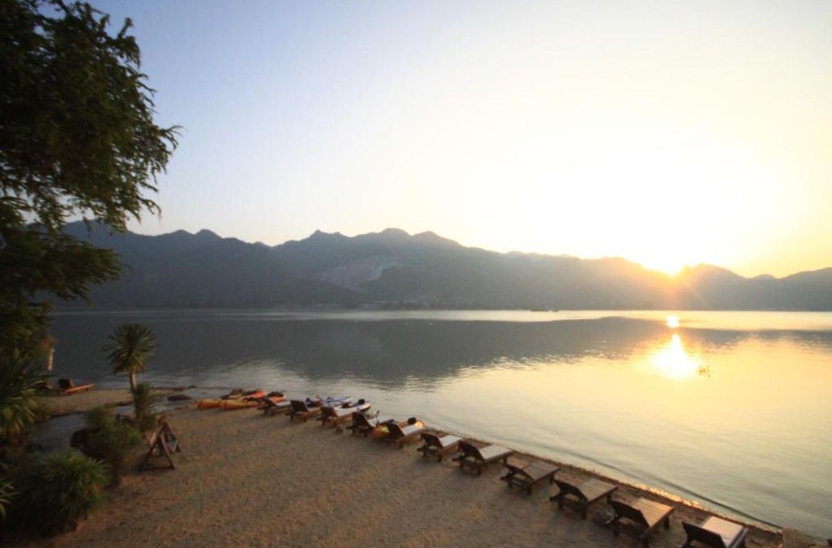 A beach with lounge chairs and a calm lake, surrounded by mountains.