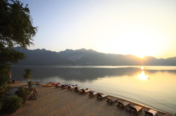 A beach with lounge chairs and a calm lake, surrounded by mountains. - Palette Monsane River Kwai Resort (Kanchanaburi)