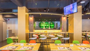 Un restaurante moderno con varias mesas y sillas, una zona de bar con estanterías verdes y un televisor de pantalla plana montado en la pared.