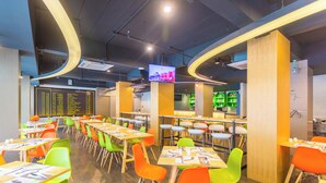 Un comedor moderno con varias mesas y sillas, un panel de llegada digital y una zona de bar con estantes verdes.