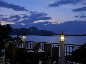 A dining area with a table and chairs, two lantern-style lights, and a view of a calm body of water with mountains in the background. - Palette Monsane River Kwai Resort (Kanchanaburi)