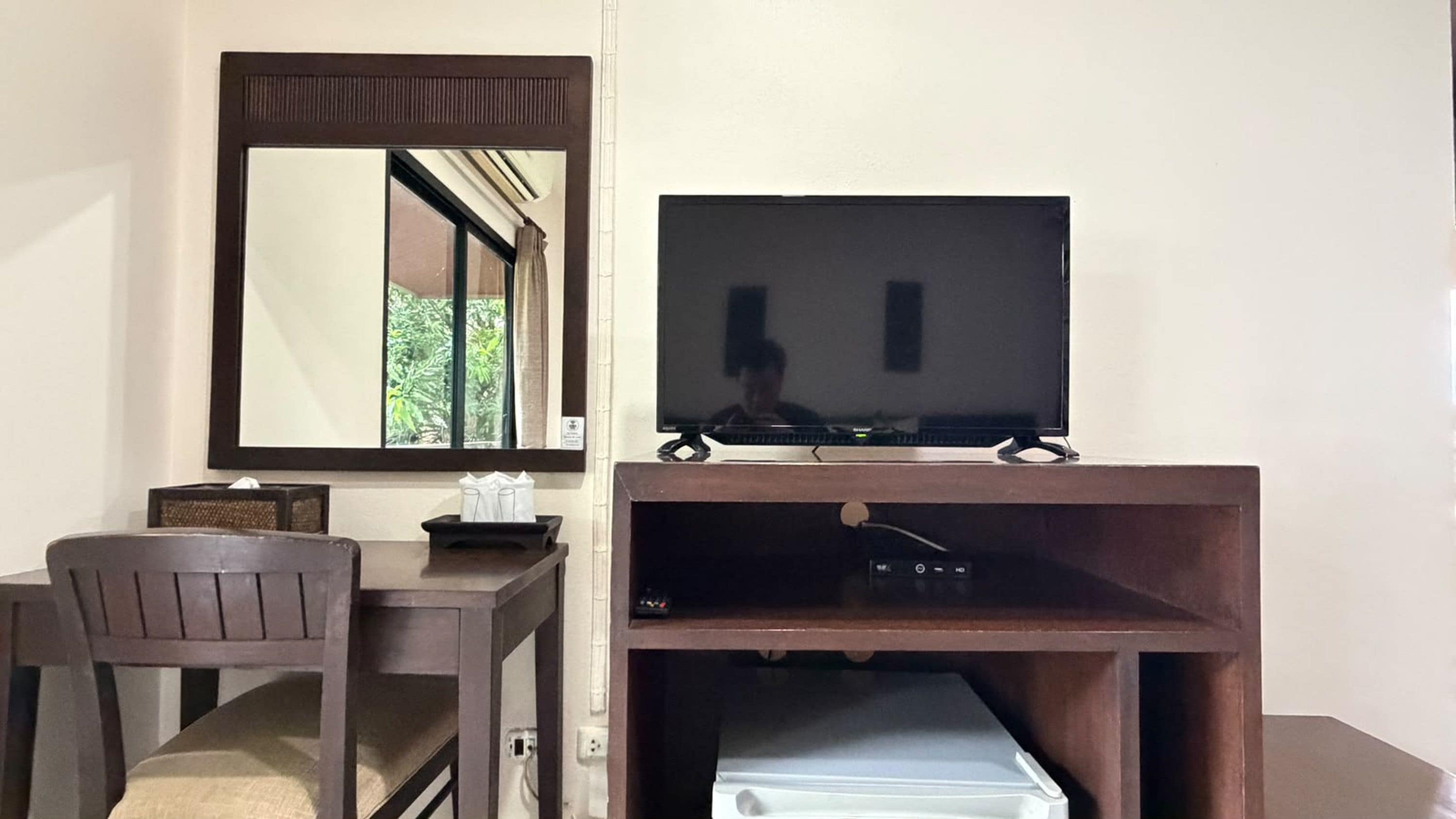 A hotel room with a wooden desk, a chair, a television on a stand, and a mirror above the desk.